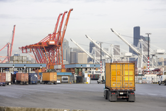 A photograph of trucks with containers entering a port gate with gantry cranes in the background.