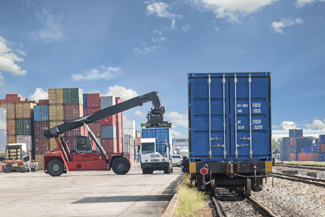 A photograph of a reach stacker, truck with container, and railcar with container with stacked containers in the background.