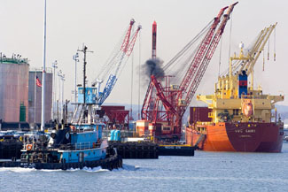 A photograph of a ship, crane, and tugboat with black smoke being emitted.