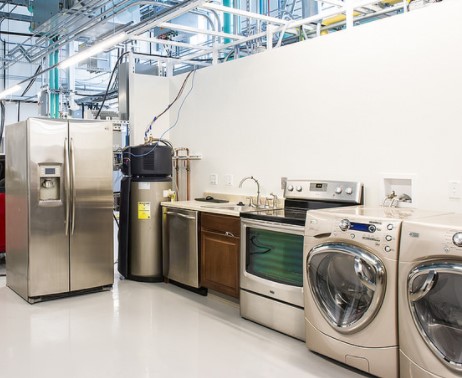 This is an image of durable goods at a store. Specifically shown are a refrigerator, washing machines and dryer, an oven with burners on top, and a kitchen sink.