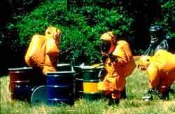 photograph of investigators in hazardous material protection suits, viewing multiple drums