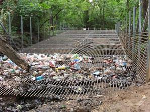 Large, metal cage-like structure with cage on bottom and two sides crossing a river and filled with trash
