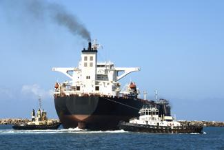 A photograph of a ship emitting black smoke and two tugboats.