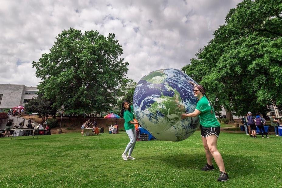 This is a picture of two women in green t-shirts holding an inflatable ball of the earth. They're outside on grass. Trees with green leaves and a cloudy sky are in the background.