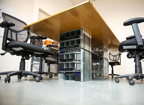 This photo shows a work space from the from the eye level of the floor. Pointing up, the shot shows chairs around a wooden table, which is supported structurally from underneath by a server chassis.