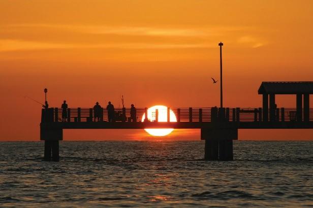 The sun sets in a vibrant orange sky, dipping below a pier and just above the line where the water meets the sky.