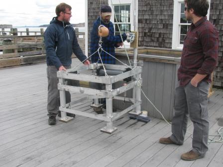 Three men stand on a deck of a pier steadying a large rack of water testing tools on an overcast day