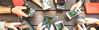 Photo of several hands holding cellphone in a circle around a table. A few purses are also present in the photo.