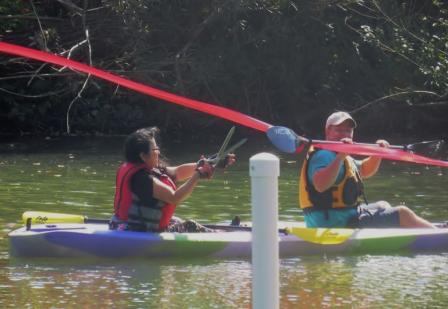 Young woman in a tandem kayak reaches out with ceremonial scissors to cut the ribbon and open the first handicap accessible boat launch in Indiana.