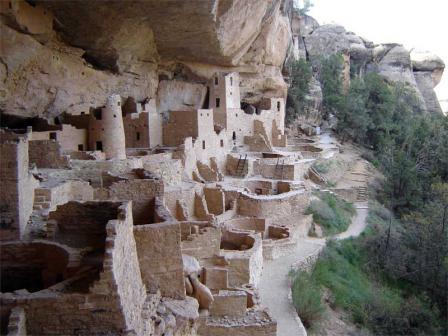 Multi-story buildings made of stone, without roofs and obviously abandoned, yet showing little sign of damage, sheltered beneath a cliff overhang.