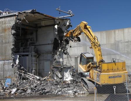 This is a picture of a partially demolished building. There also is debris on the ground in front of the part of the building that is demolished, and a yellow crane is picking up some debris.