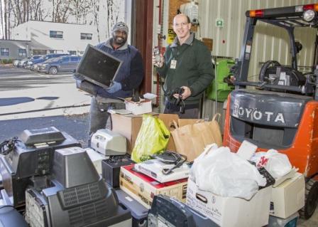 This is a picture of two gentlemen holding different types of e-waste. The guy on the left is smiling and holding a laptop while the guy on the right is smiling and holding a phone and cord. There are boxes of e-waste on the ground in front of them.