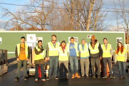 This is a picture of volunteers standing in a horizontal line, posing for a picture. They're wear lime green reflecting vests and standing in front of a green, one-story building and leafless trees.