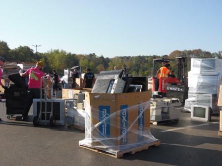 This photo shows materials on palettes, a furniture dolly and a forklift. The materials are outside in a parking lot. Trees and a clear buy sky are in the background.