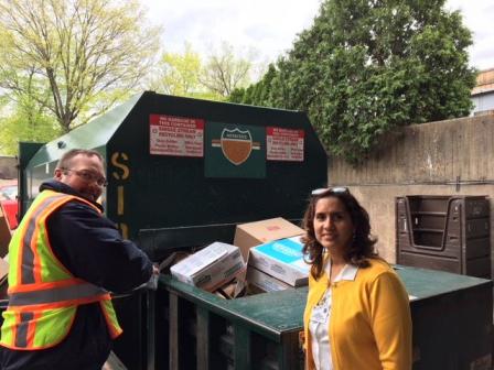 This is a picture of a man and a woman posing in front of a green recycling bin. It has cardboard in it.