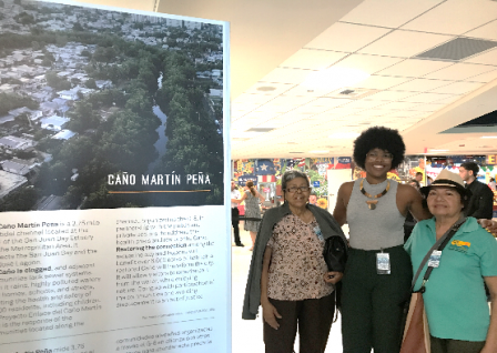 Three women pose in front of San Juan Bay Estuary Exhibition at the San Juan International Airport