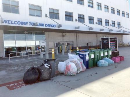 Two large black plastic bags and gray plastic bins outside filled trash and about seven large clear plastic bags filled with cans and cardboard and six large blue and green plastic compost and recycling bins placed along the airport curb for collection.