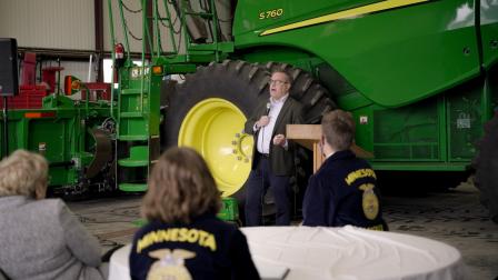 photo of Wheeler spaking to an audience and a large green tractor