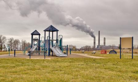 A plume of emissions rises from a factory smokestack near an empty playground