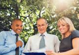 Two men and one woman in a park with trees read from a clipboard